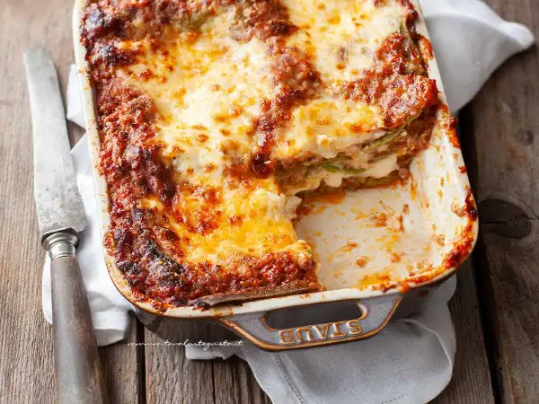 Lasagne alla bolognese appena sfornate, dorate e filanti, in una teglia Staub. Un coltello vintage è appoggiato accanto.