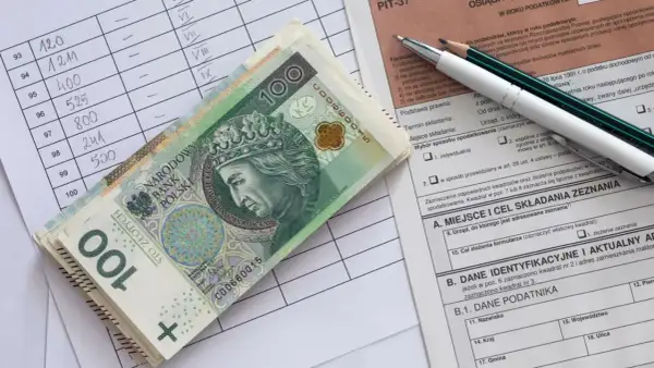 Stos banknotów 100 zł leży na formularzu PIT. Obok arkusz z danymi i ołówki.