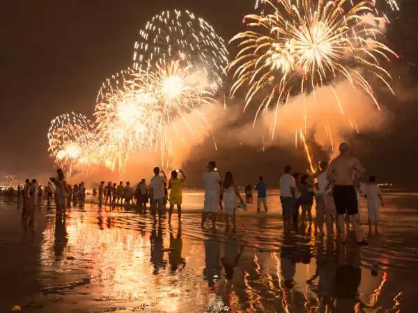 Ludzie na plaży podziwiają spektakularny pokaz fajerwerków. To idealny sylwester w ciepłych krajach, z odbiciami świateł w wodzie.
