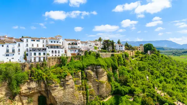Vita hus i Ronda, Spanien, klättrar uppför en grönskande klippa under en blå himmel med moln.