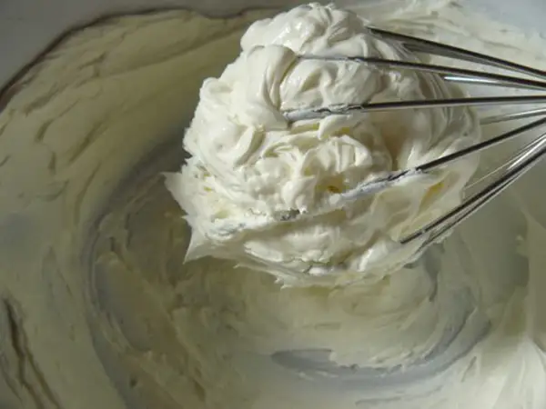 Un fouet de cuisine retient une généreuse portion de glacage mascarpone crémeux, prêt à garnir un gâteau.
