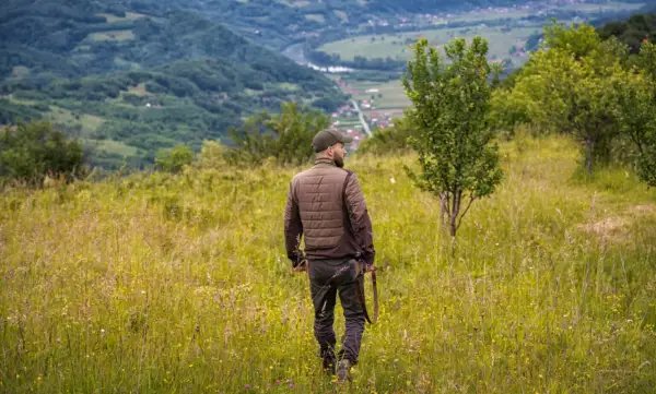 Uomo con fucile attraversa prati fioriti, godendosi la vista dei passi di montagna e della valle sottostante.