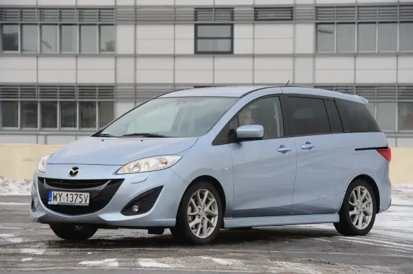 Jasnoniebieska Mazda 5 z nowymi żarówkami w reflektorach, zaparkowana przed nowoczesnym budynkiem.