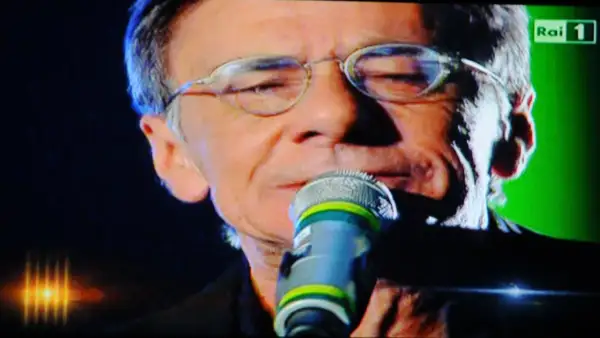 Roberto Vecchioni canta "Chiamami ancora amore" con passione, il microfono vicino alle labbra, sullo sfondo verde di Rai 1.