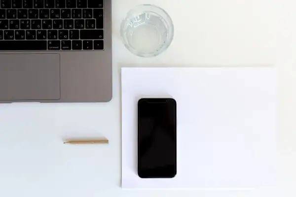 Przygotowanie do naklejania szkła na telefon. Obok laptopa i szklanki wody leży czarny smartfon na białej kartce.