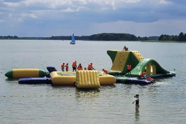 Rodzinna zabawa na wodnym placu zabaw na Mazurach. Dzieci w kamizelkach ratunkowych wspinają się i ślizgają po dmuchanych konstrukcjach.
