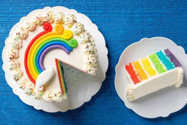 Torta arcobaleno con decorazioni colorate e una fetta servita su un piatto.