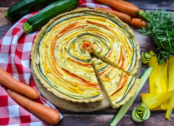 Bunte quiche vegetarisch mit Zucchini und Karotten, kunstvoll im Kreis arrangiert. Daneben frisches Gemüse.