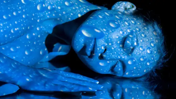 Una persona con la piel pintada de azul, cubierta de gotas blancas que parecen reflejar **que es la luz azul**.