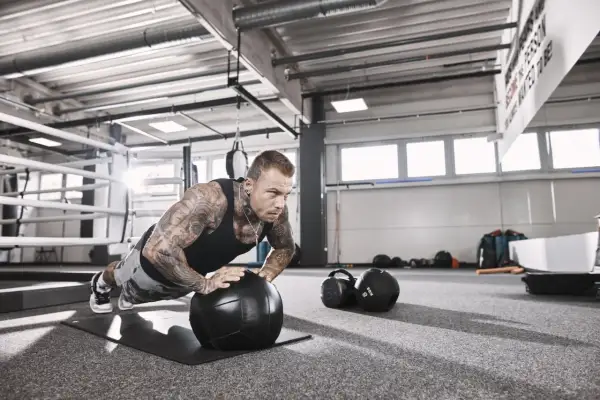 Mann mit Tattoos macht Liegestütze auf einem Medizinball, fokussiert auf sein hypertrophie training. Im Hintergrund ein Boxring und Trainingsgeräte.