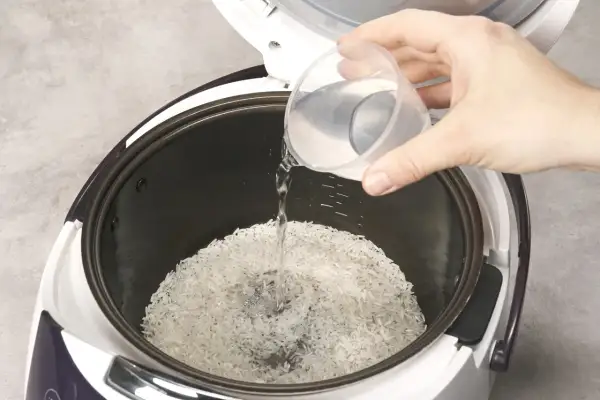 Beim Reis kochen wie viel Wasser? Eine Hand gießt Wasser in den Reiskocher über den Reis.