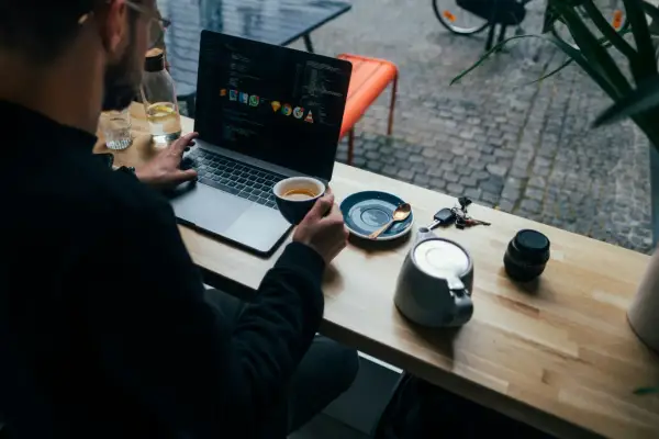 Mężczyzna pracuje na laptopie, popijając kawę. Widać kod na ekranie, klucze i aparat. To przykład multitasking co to znaczy być produktywnym.