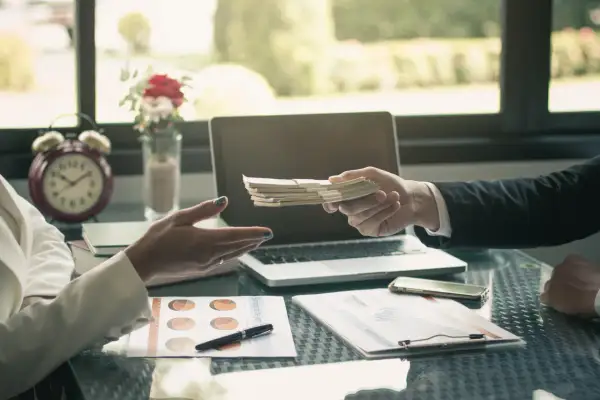 Dłoń przekazuje plik banknotów, co stanowi potwierdzenie przyjęcia gotówki. Na stole leżą dokumenty, laptop i telefon.