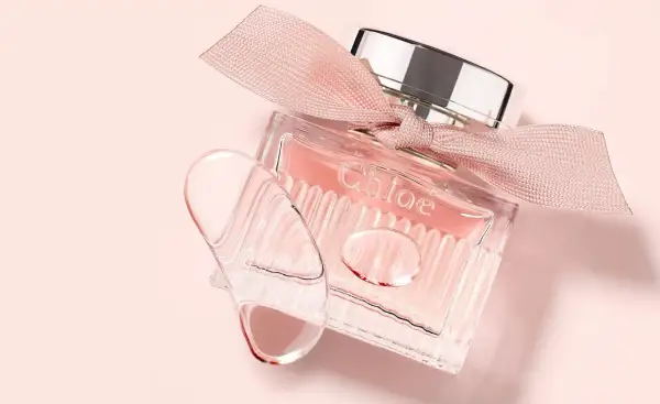Szklana buteleczka L'eau de Chloe z różową wstążką i kroplami perfum na delikatnym, różowym tle.