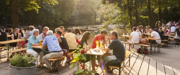 Menschen genießen das Leben im cafe am neuen see. Sie sitzen an Holztischen, trinken Bier und unterhalten sich. Im Hintergrund fließt ein ruhiger Fluss.