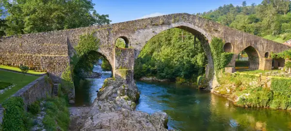 El puente del Sella, de piedra y cubierto de vegetación, cruza un río de aguas verdes rodeado de árboles frondosos.