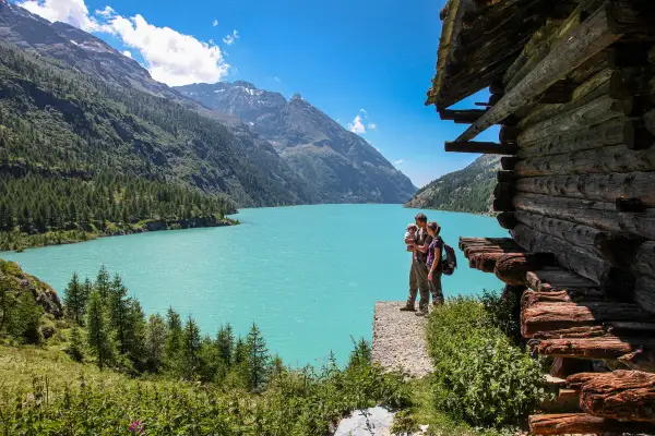 Un couple avec un bébé admirent le lac d'Aoste, entouré de montagnes verdoyantes et d'une cabane en bois rustique.