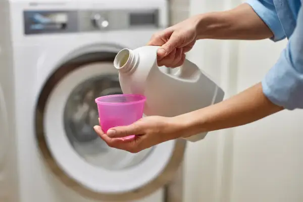 Mani versano candeggina in un misurino rosa, vicino a una lavatrice. Quanto tempo ammollo candeggina?