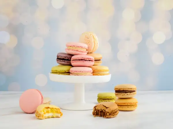 Bunte Macarons auf einem weißen Tortenständer, einige angeschnitten, vor unscharfem Bokeh-Hintergrund.