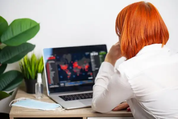 Kobieta z rudymi włosami patrzy na ekran laptopa z mapą świata i danymi. Obok leży maseczka i płyn do dezynfekcji.