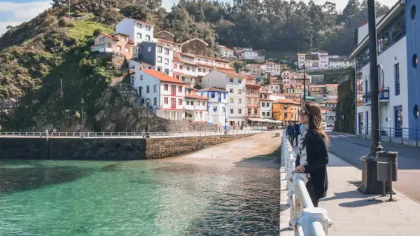 Una mujer admira la costa de **pueblos de Asturias cerca de Galicia**. Casas coloridas se aferran a la ladera verde junto a un mar turquesa.