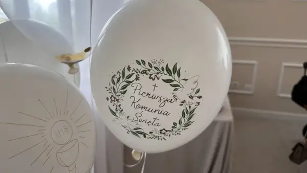 Białe balony z napisem "Pierwsza Komunia Święta" i motywem gołębicy oraz kielicha z hostią.