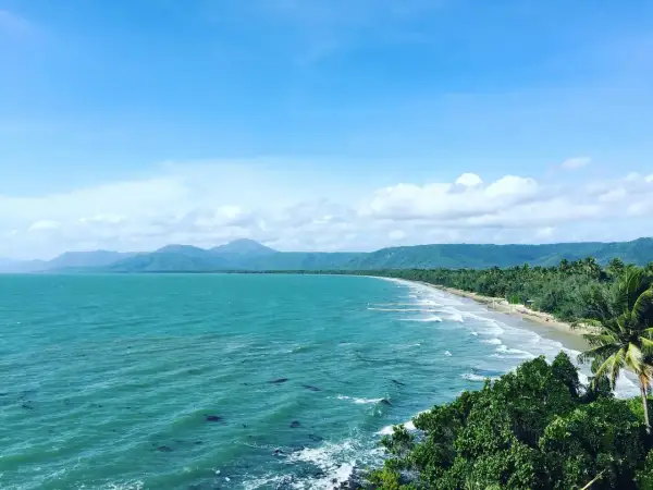 Vacker kustlinje vid Port Douglas med turkost hav, grönskande berg och en gyllene strand under en molnig himmel.