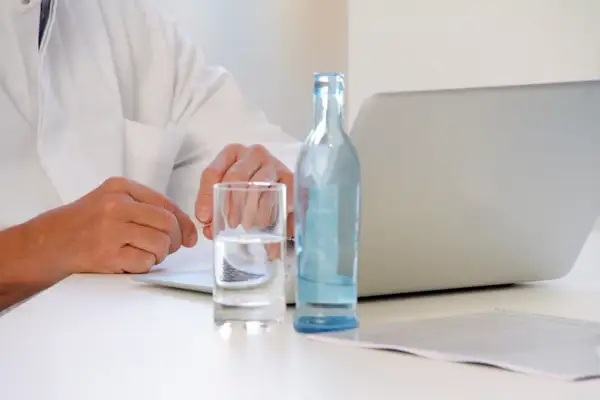 Lekarz w białym kitlu przy laptopie, obok szklanka wody i butelka. Czy można pić wodę przed tomografią z kontrastem? Zapytaj lekarza.