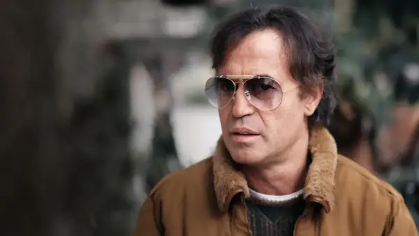Close-up of a man with dark hair, wearing aviator sunglasses and a brown jacket with a fur collar.