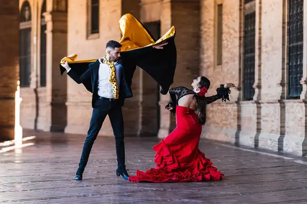Pareja bailando flamenco con pasión. Él, con capa negra y dorada, ella, con vestido rojo voluminoso. Puro jaleo flamenco.