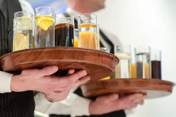 Kelnerzy niosą tace z drinkami, idealne na dzień wódki. W szklankach widać wodę z cytryną, colę i sok pomarańczowy.