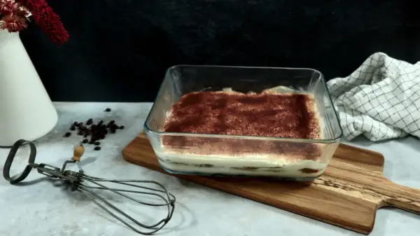 Un délicieux tiramisu 6 personnes, prêt à être dégusté, saupoudré de cacao. Un fouet ancien et des grains de café complètent la scène.