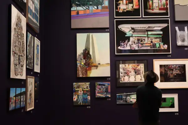 A man in a hat views diverse artworks in a gallery, showcasing vibrant cityscapes and street scenes, typical of young art galleries.