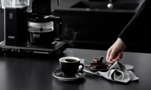 En hand sträcker sig efter chokladbitar bredvid en kopp rykande kaffe. Kaffebryggaren i bakgrunden ser ut att frysa kaffe för en perfekt start på dagen.