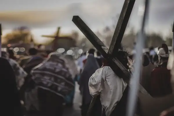 Osoba z koroną cierniową niesie krzyż, idąc w procesji. W tle widać inne krzyże i ludzi, przypominające stacje drogi krzyżowej.
