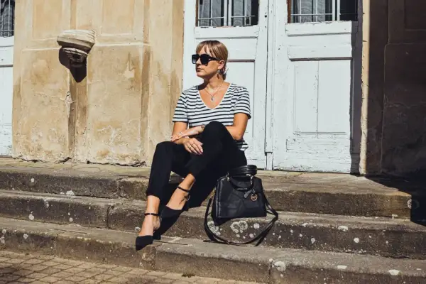 Elegancka kobieta w czarnych cygaretkach i bluzce w paski, siedząca na schodach. Stylowa torebka Michael Kors dopełnia całość.
