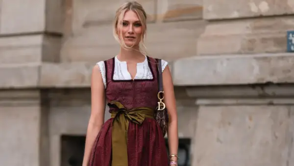 Blonde Frau in einem roten Dirndl mit weißer Bluse und einer grünen Schürze. Sie trägt eine Dior-Tasche.
