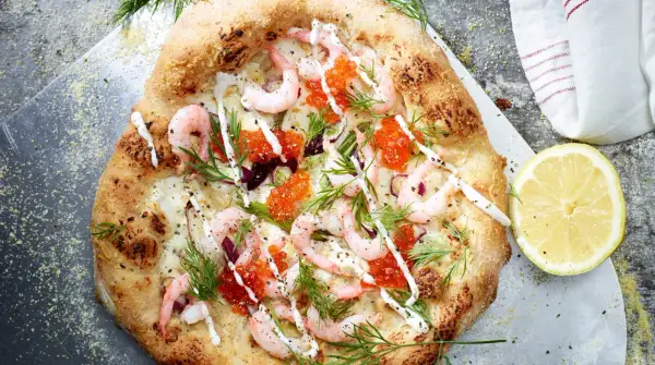 En läcker pizza med räkor och mozzarella, toppad med rom och dill. En citronskiva ligger bredvid.