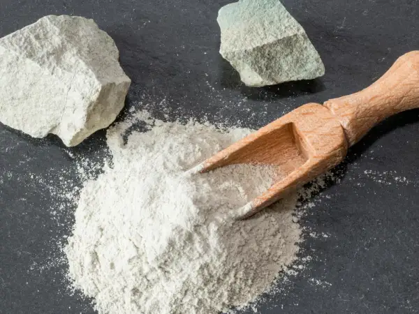 Feiner Pulver aus Magnesium-Aluminium-Silikat wird mit einer Holzschaufel auf dunklem Stein verteilt, daneben liegen zwei Brocken des Materials.