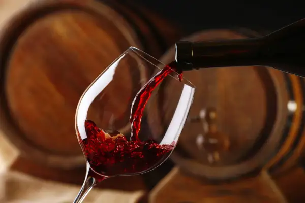 Czerwone wino leje się do kieliszka, a w tle widać drewniane beczki, które opowiadają historię wina.