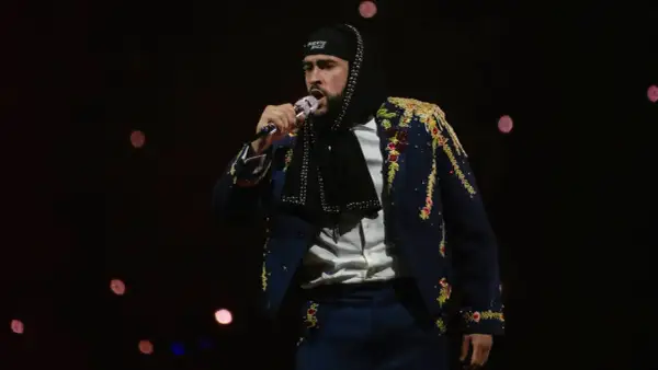 Bad Bunny w ozdobnej marynarce śpiewa na koncercie na ciemnym tle z rozmazanymi światłami.