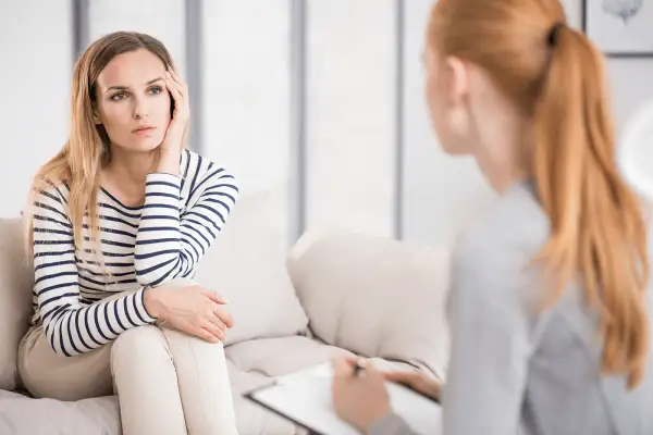 Kobieta w paski siedzi na kanapie, zamyślona, podczas rozmowy z terapeutką. Czy psycholog pomaga? Tak, widać, że wsparcie jest potrzebne.