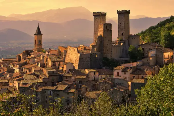 Borghi da visitare in Abruzzo: un castello medievale domina un villaggio arroccato, con torri imponenti e case in pietra sotto un cielo dorato.