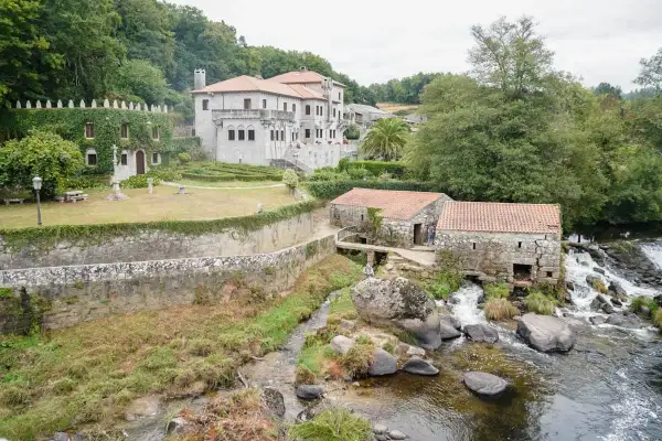 Antigua barcala con molinos de piedra junto a un río caudaloso, con una casa señorial cubierta de hiedra al fondo.