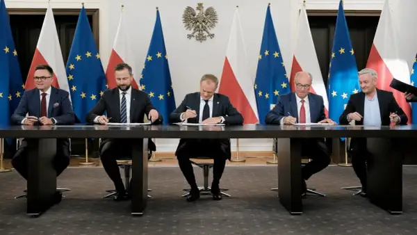 Pięciu mężczyzn w garniturach siedzi przy stole, podpisując dokumenty. W tle flagi Polski i UE. Może to być ważny moment dla sejmowej komisji śledczej.