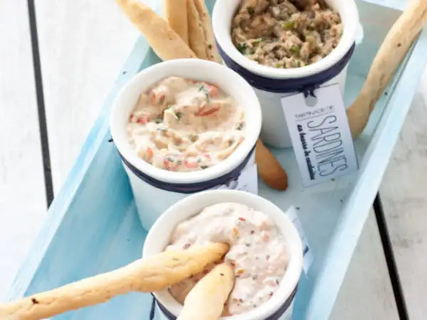 Trois ramequins de rillettes de la mer, dont une tartinade de sardines, présentés sur un plateau bleu avec des gressins.