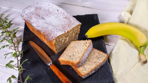 Ein saftiger Bananenkuchen, bestäubt mit Puderzucker, liegt auf einem schwarzen Brett. Daneben zwei Scheiben und eine reife Banane. Das ist der beste Bananenkuchen der Welt!