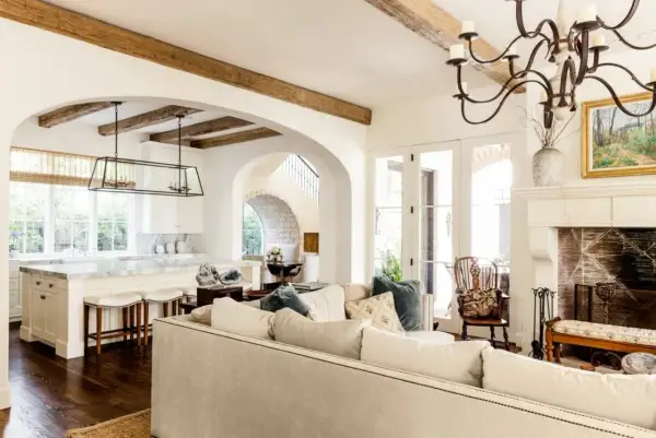 A cozy living room with a fireplace and kitchen, featuring exposed wooden beams and arched doorways, embodying a warm mediterranean interior design.