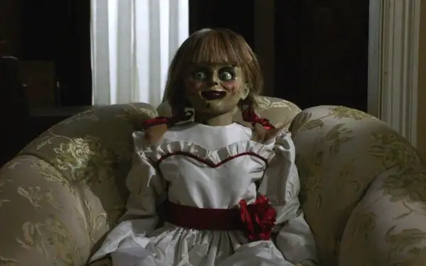 Den läskiga riktiga Annabelle dockan sitter i en blommig soffa, med sina stora ögon och röda rosett.