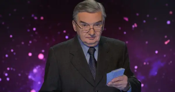 Tadeusz Sznuk w garniturze i okularach trzyma w ręku niebieskie karty na tle fioletowej, rozmytej scenografii.